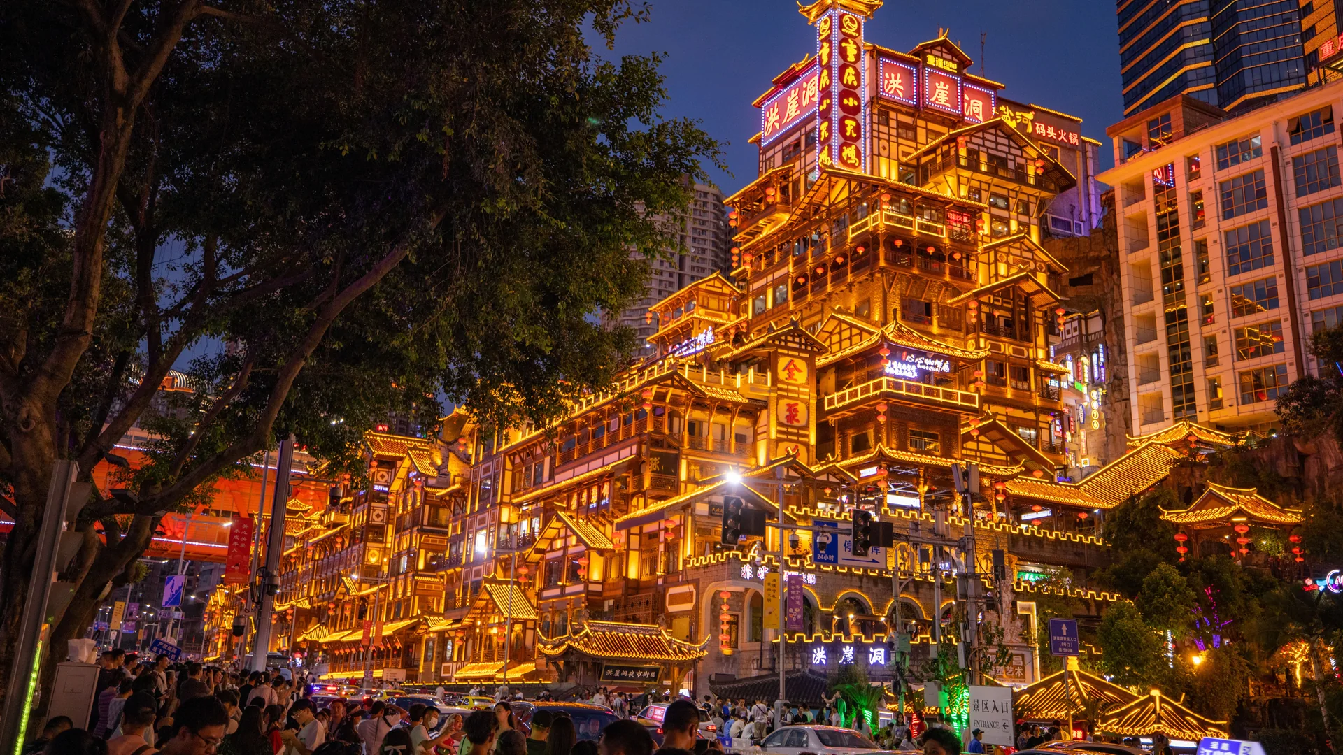 Hongya Cave, the stilt-style riverfront complex in central Chongqing, lit gold at dusk with a tree in the foreground. It is a different place from the theatre.