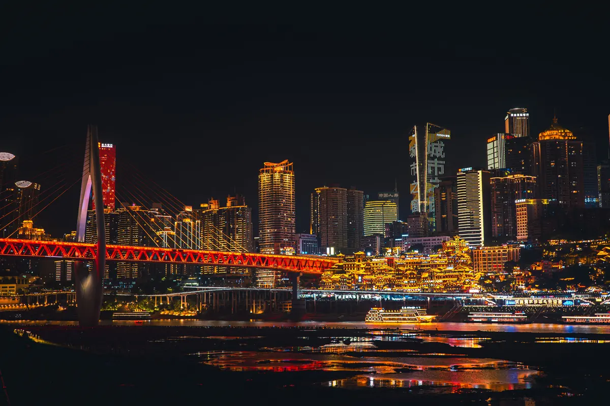 Chongqing's skyline across the river at night, with a red cable-stayed bridge and the gold-lit riverside complex.