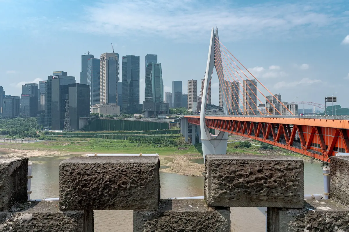 A cable-stayed bridge with a red deck crossing the river in Chongqing, with towers behind it, seen between stone battlements